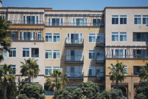 Modern apartment building with balconies and large windows under a clear blue sky. Palm trees in front create a sunny, relaxed atmosphere.